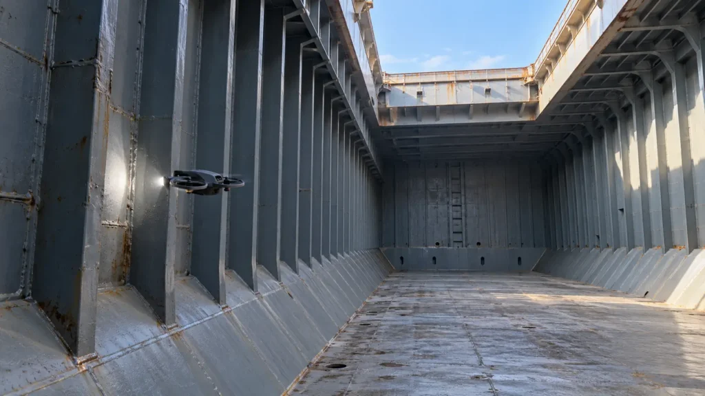 Drohne mit Zusatzlicht vor einer Stahlwand im leeren Schiffsladeraum mit sichtbaren Spanten und offener Ladeluke.